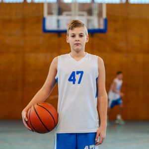 Portrait of a young basketball player standing on court with a basketball in his hands and looking at the camera. A basketball kid posing on court with a ball. In background is his team on training.