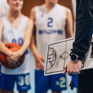 Cropped picture of a basketball trainer with clipboard standing at indoor court with his junior team and working on game strategy. Close up of basketball coach's hand holding clipboard on training.